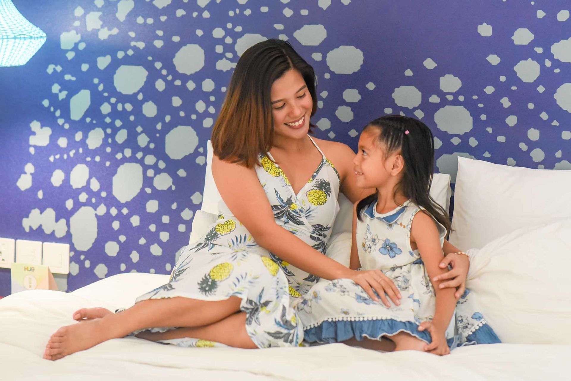 Woman and child in matching dresses sitting on a bed in front of a patterned wall, smiling at each other.
