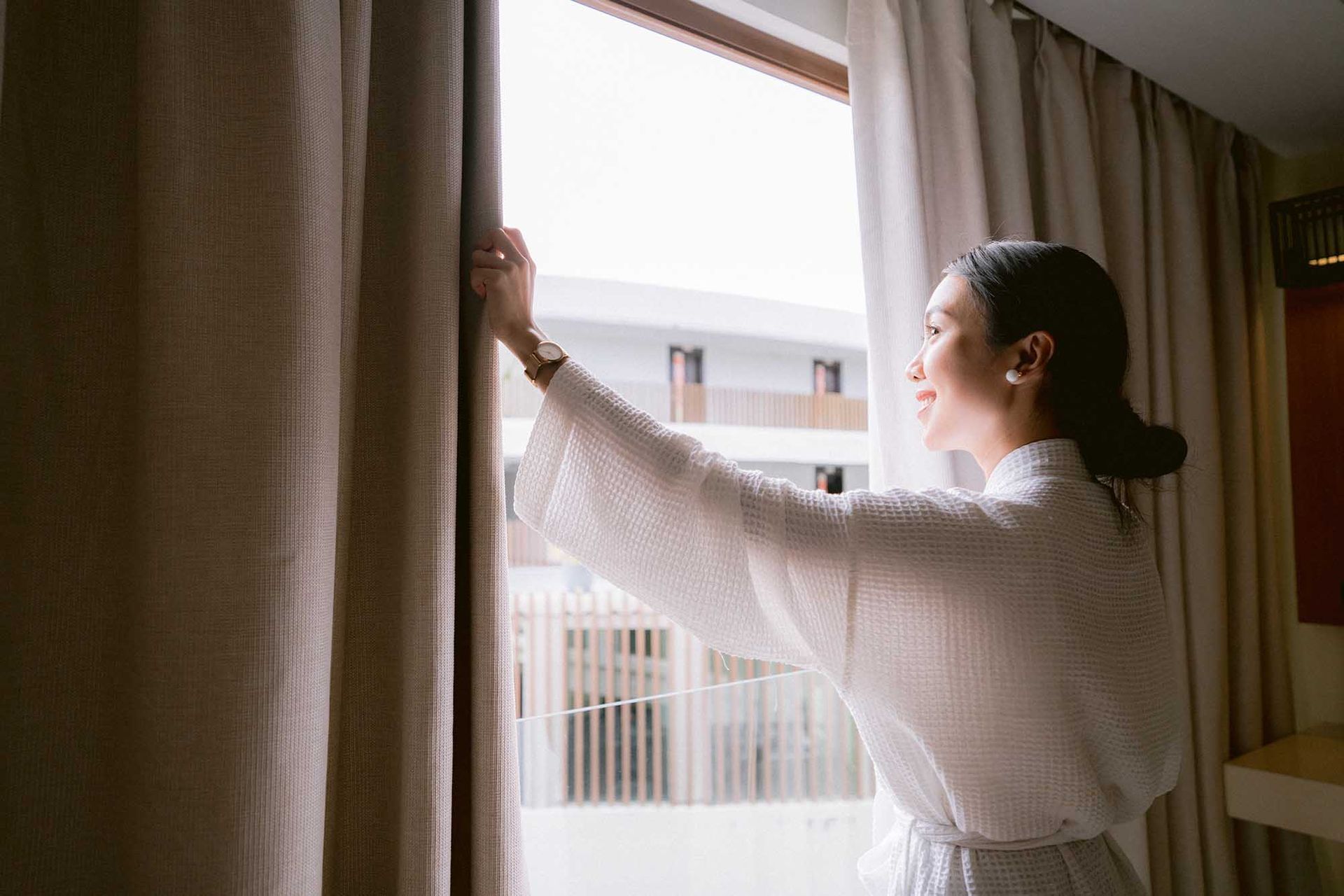 Woman in robe opening curtains to look out the window.