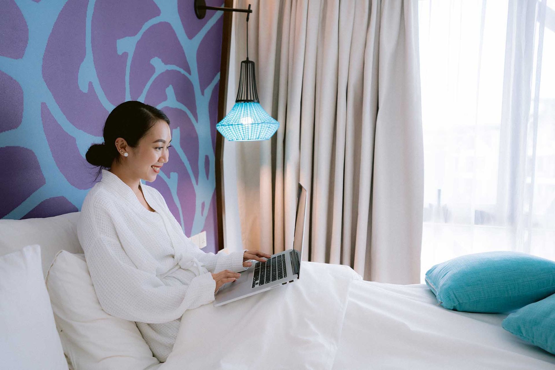 Person in a white robe using a laptop in a bright bedroom with teal pillows and sheer curtains.
