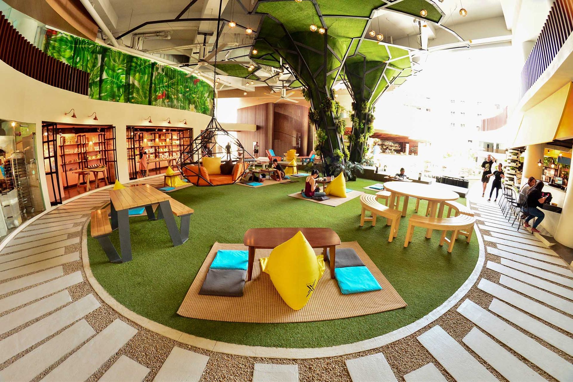 Indoor recreation area with artificial grass, seating, floor cushions, and decorative trees under high ceilings.