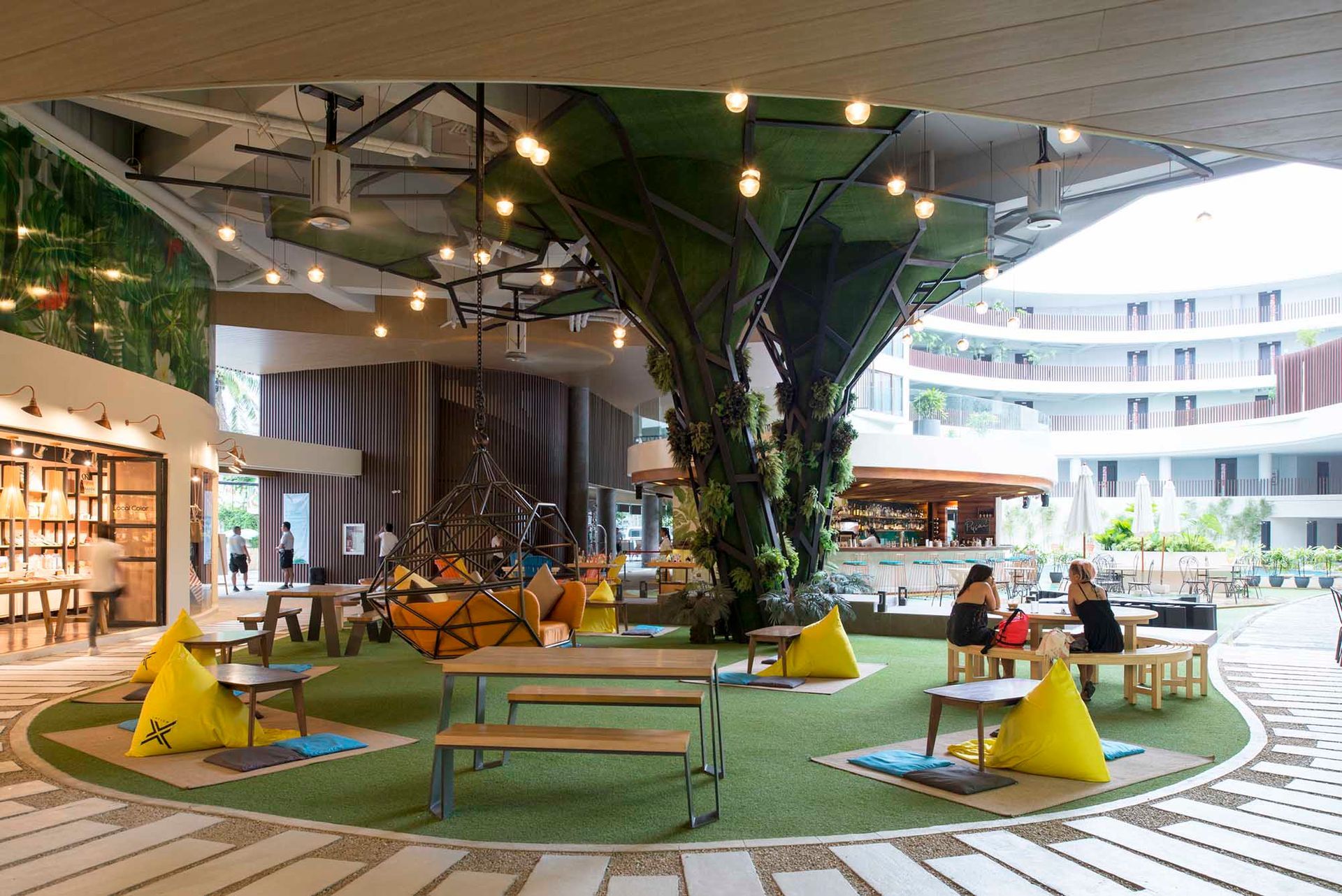 A modern, open-air resort lobby with artificial grass, yellow beanbag chairs, picnic tables, and a large central tree.