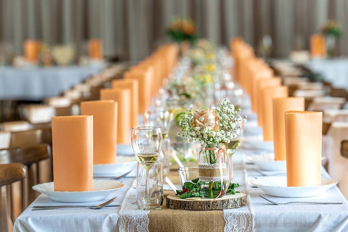 Long dining table set for a wedding, featuring orange napkins, flower centerpieces, and place settings.