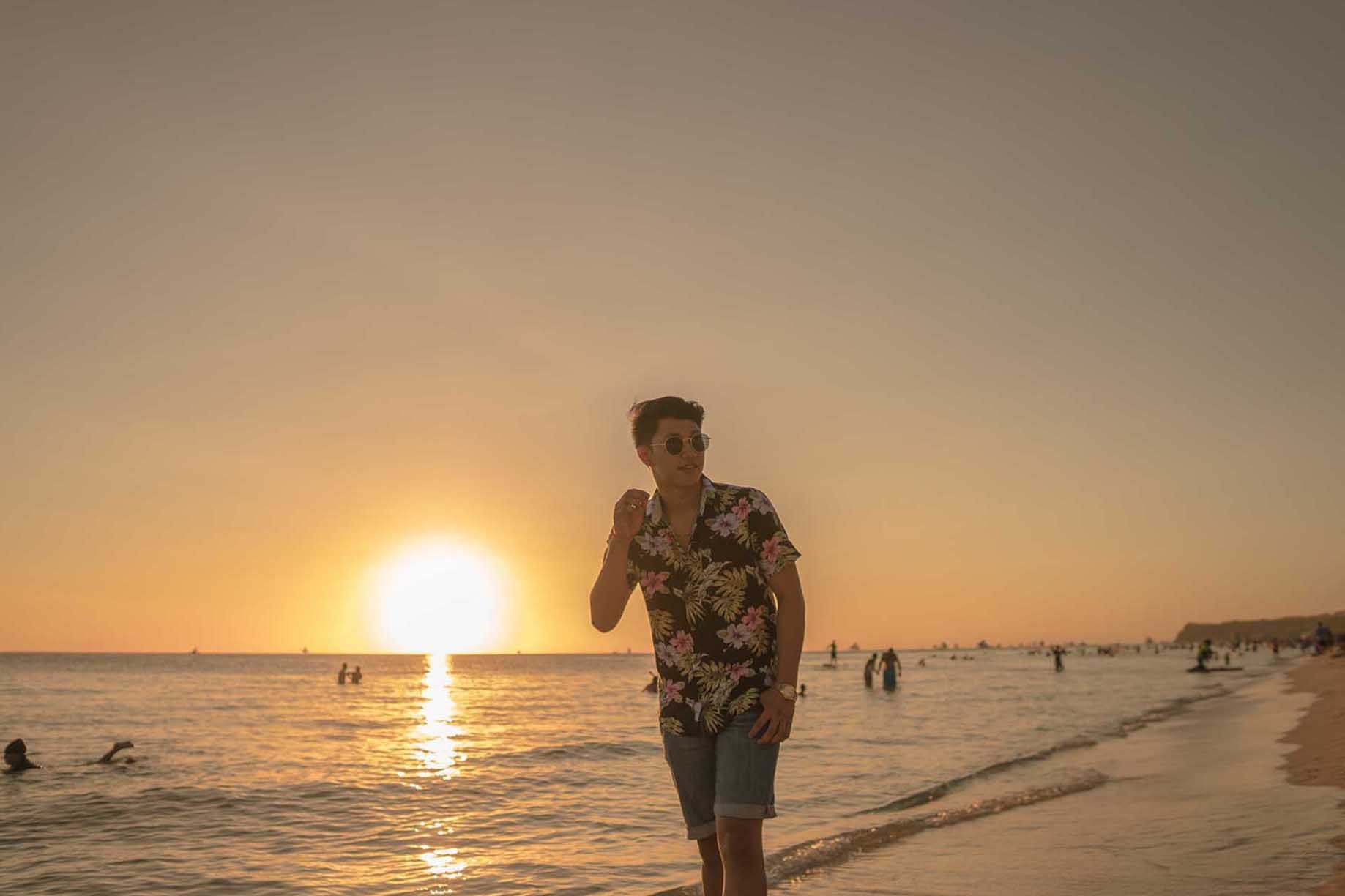 Man in floral shirt walks along beach at sunset, waves lapping.
