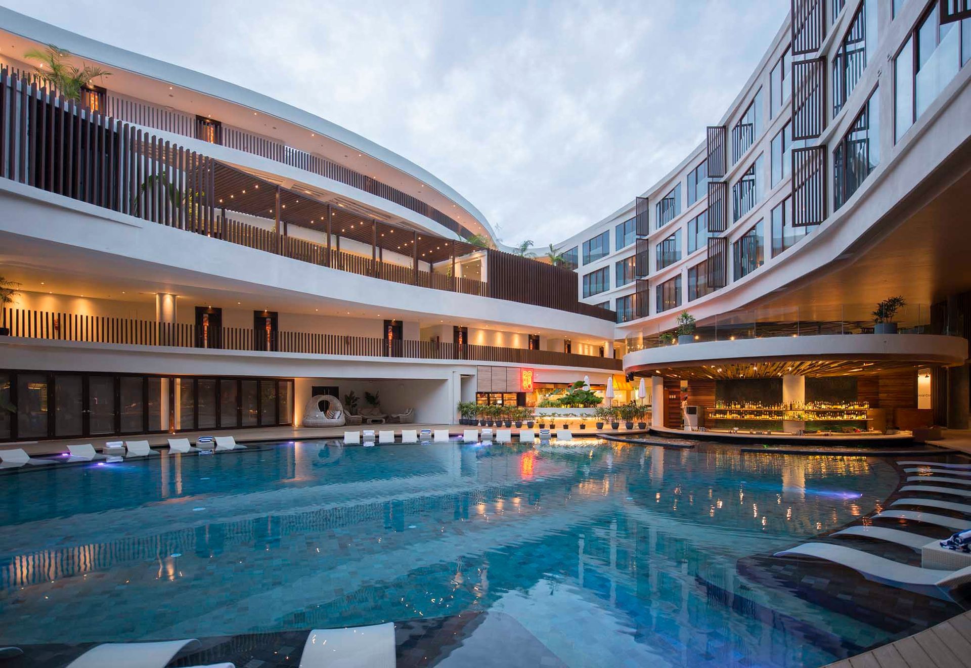 Exterior view of a modern hotel with a pool. Buildings surround the pool. Cloudy sky.