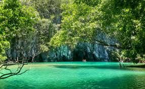 Lush green trees frame turquoise water leading to a cave opening in a rocky cliff face.