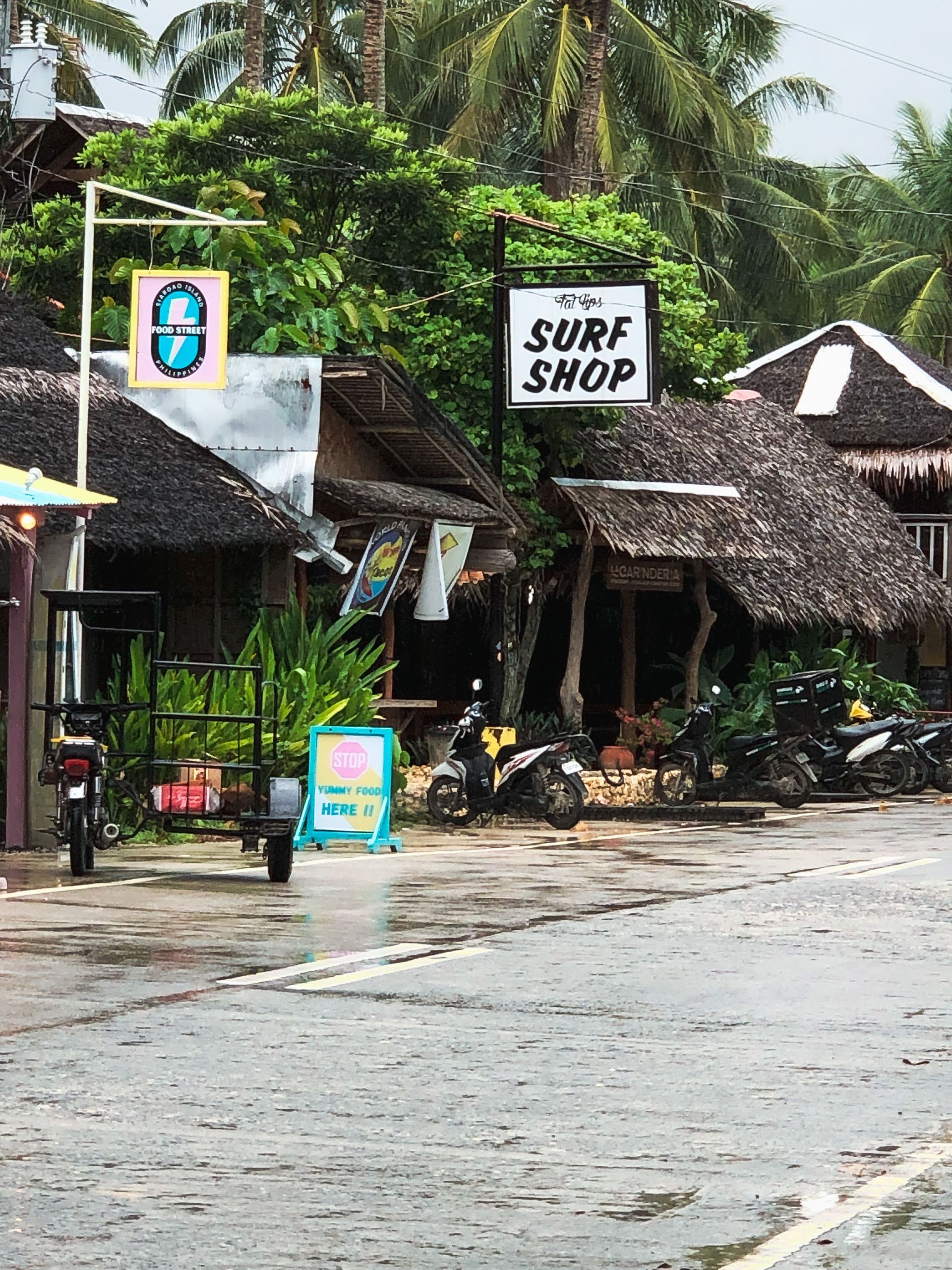 Rainy beachside surf shop with thatched huts and palm trees along a wet street