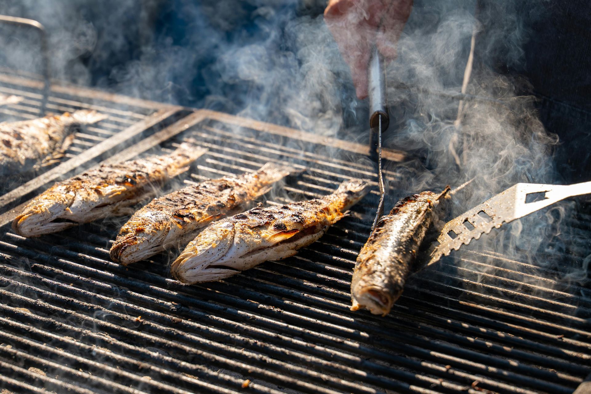 Grilled whole fish on a barbecue, person flipping a fish with a spatula, smoke rising.