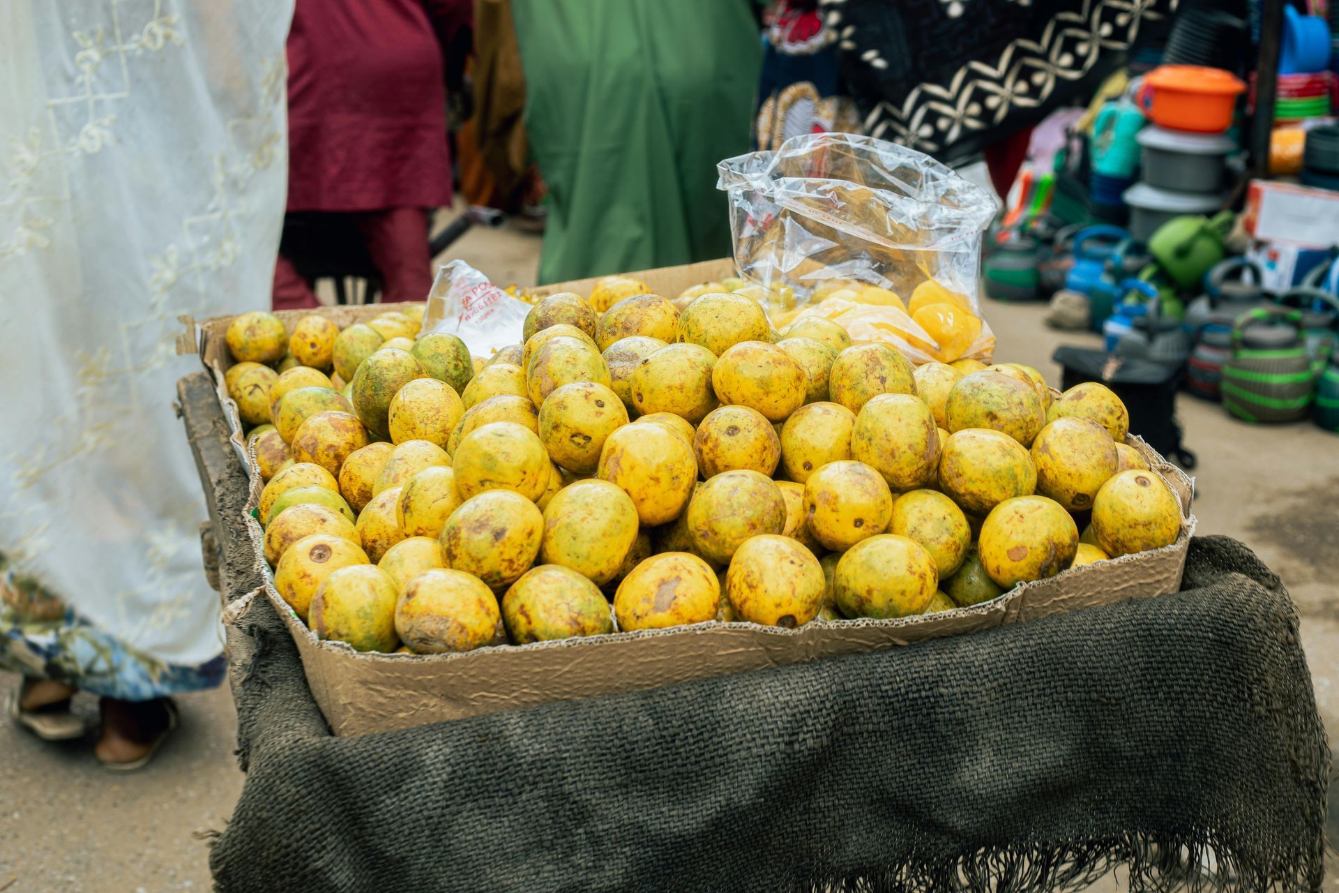 Box of yellow mangoes for sale at an outdoor market, with people in the background.