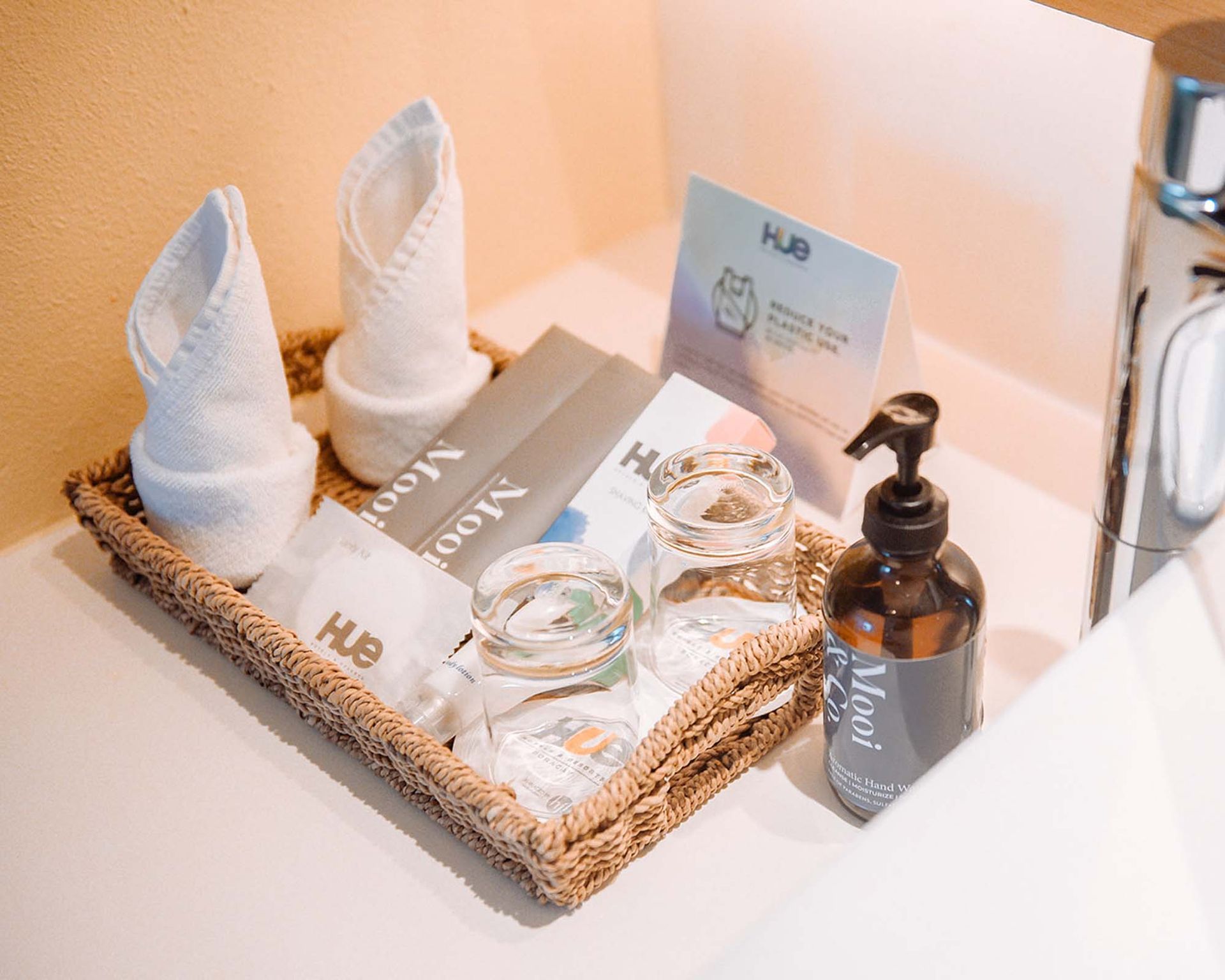 A wicker basket on a counter holds folded white towels, toiletries, glasses, and a soap dispenser in a bathroom.