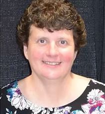 Woman with short brown curly hair smiles, wearing a floral top.
