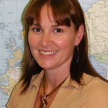 Woman with brown hair smiling, wearing tan shirt and necklace, in front of a map.