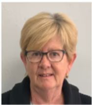 Woman with blonde hair and glasses smiles, wearing a black jacket, indoors.