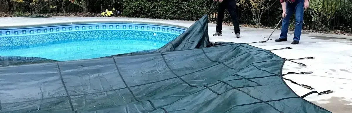 Two people stand on a pool deck, partially pulling a large, dark green mesh safety cover over an outdoor swimming pool.