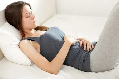 Woman lying on a bed, clutching her abdomen, possibly in pain. Wearing gray clothes, white bedding.