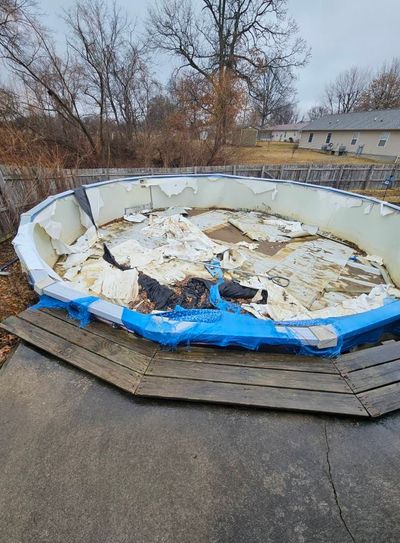 An above-ground pool in a backyard with a damaged blue cover and liner in a fenced-in, overcast outdoor setting.