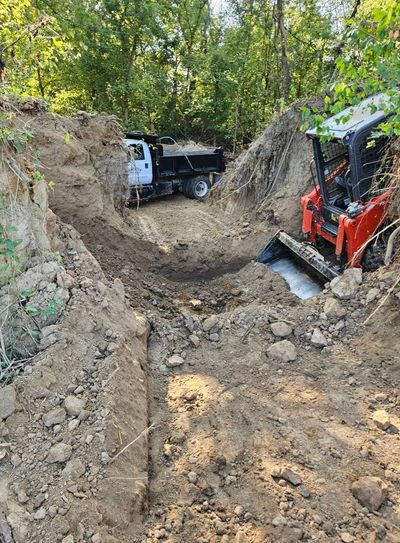 A white dump truck and a red skid steer loader parked in a deep, excavated dirt trench surrounded by trees.