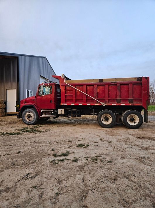 A red dump truck parked on a gravel lot next to a large metal storage building under a clear sky.
