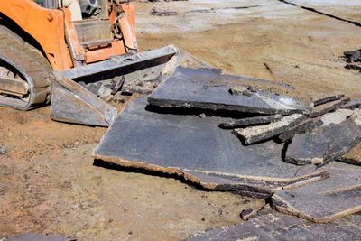 An orange construction vehicle sits on dirt next to slabs of broken asphalt, demonstrating road removal and demolition.