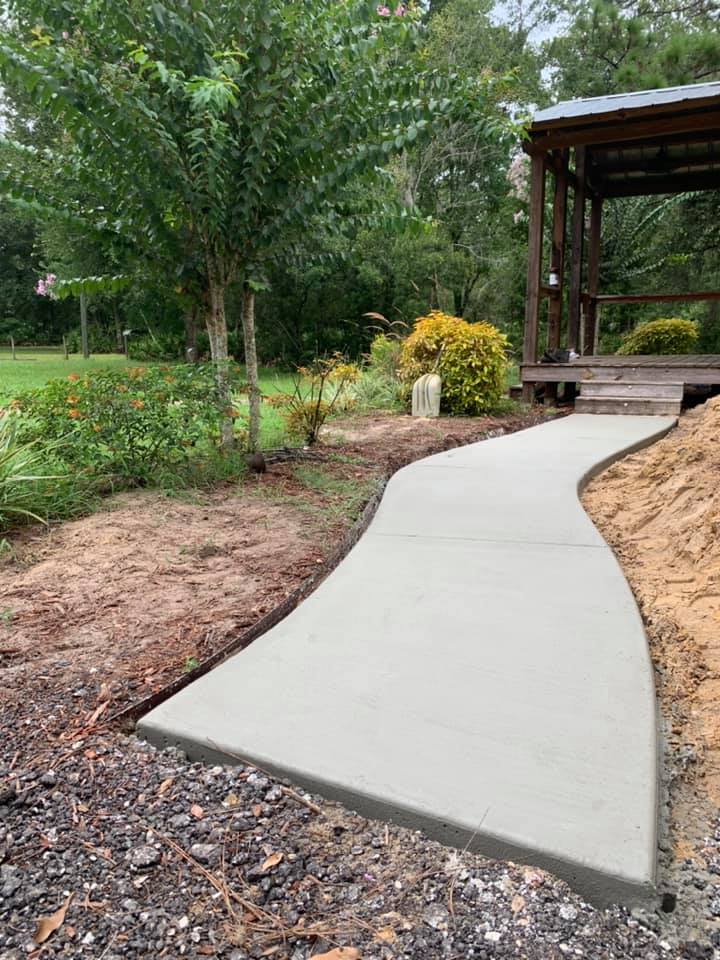 Concrete sidewalk curves through a yard, leading to a wooden structure. Trees and flowers border the path.