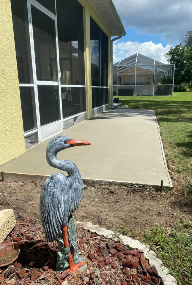 A concrete patio outside a yellow house with a heron statue, screened porch, and grassy yard.