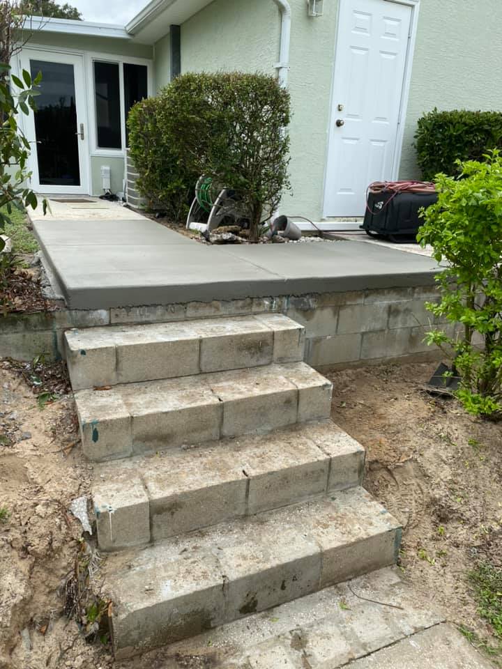 Concrete steps and platform leading to a house with a white door and glass doors. Green bushes are on either side.