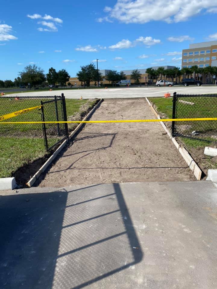 Dirt pathway blocked by yellow tape and black fence. Concrete borders edge the path. Sunny outdoor setting.