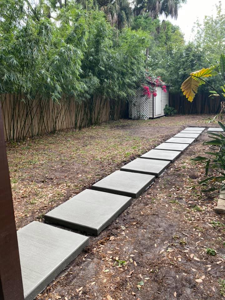 A concrete pathway leads through a yard with dirt, a shed with pink flowers, and bamboo trees.