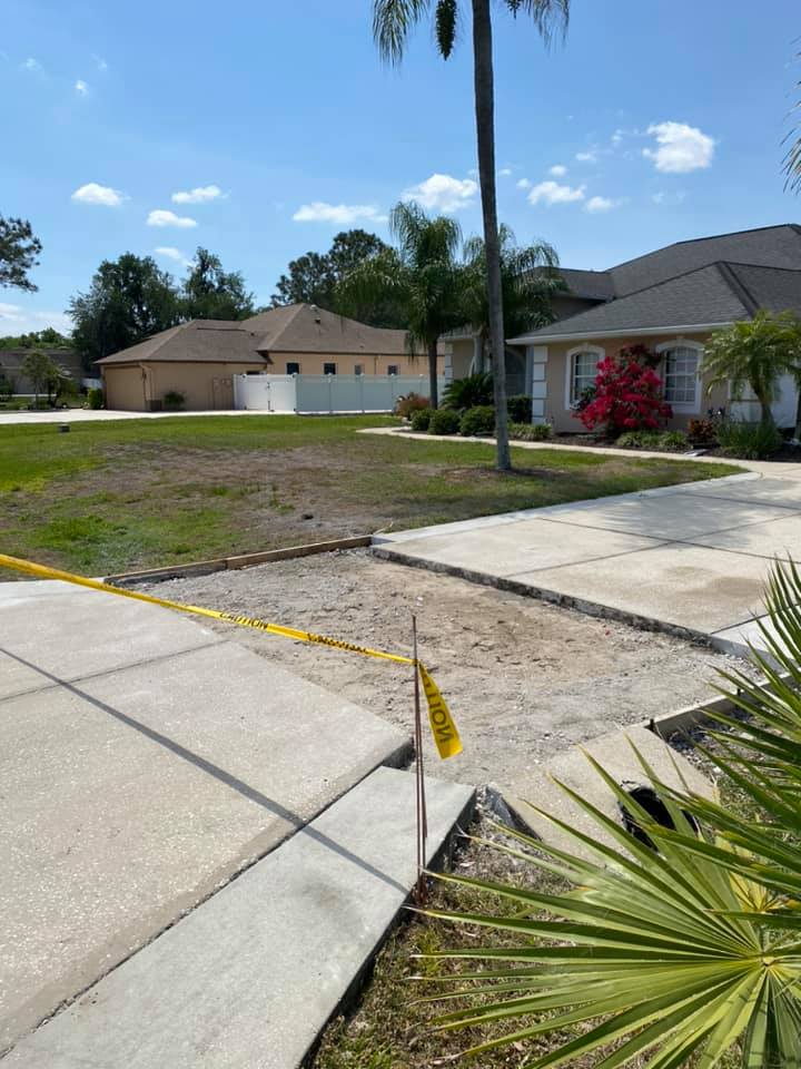 Driveway construction site with caution tape, gravel, and partial concrete paving. Sunny day, residential area.