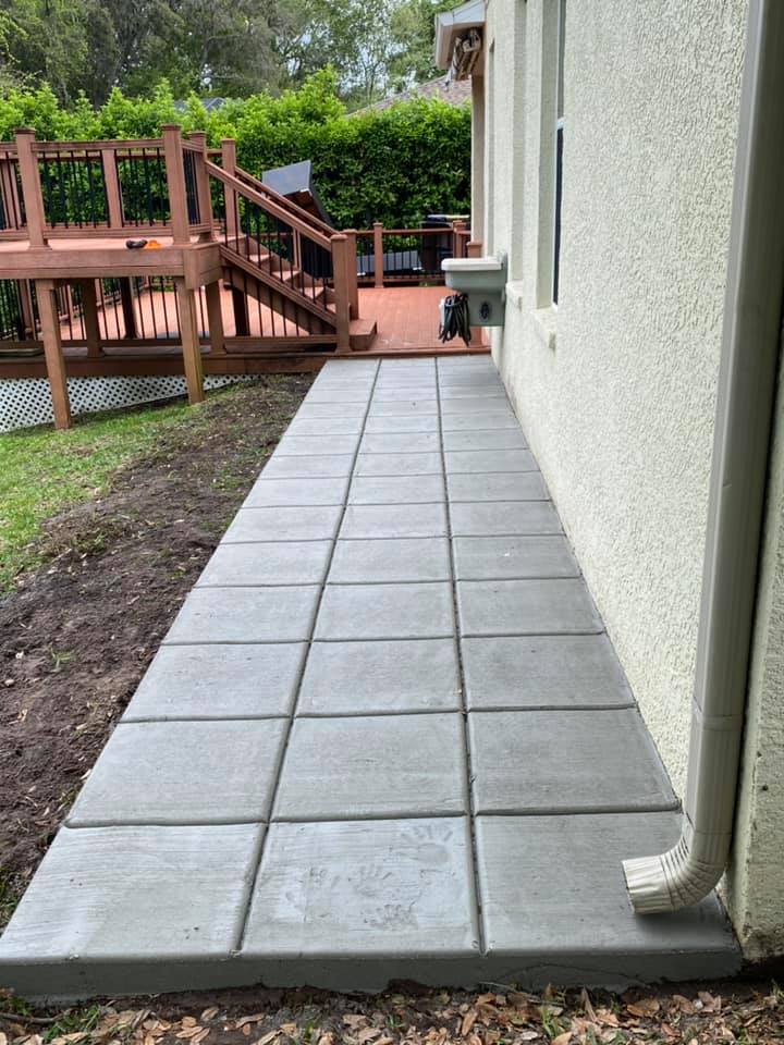 Concrete paver walkway next to a light beige house and a wooden deck, with green grass on either side.