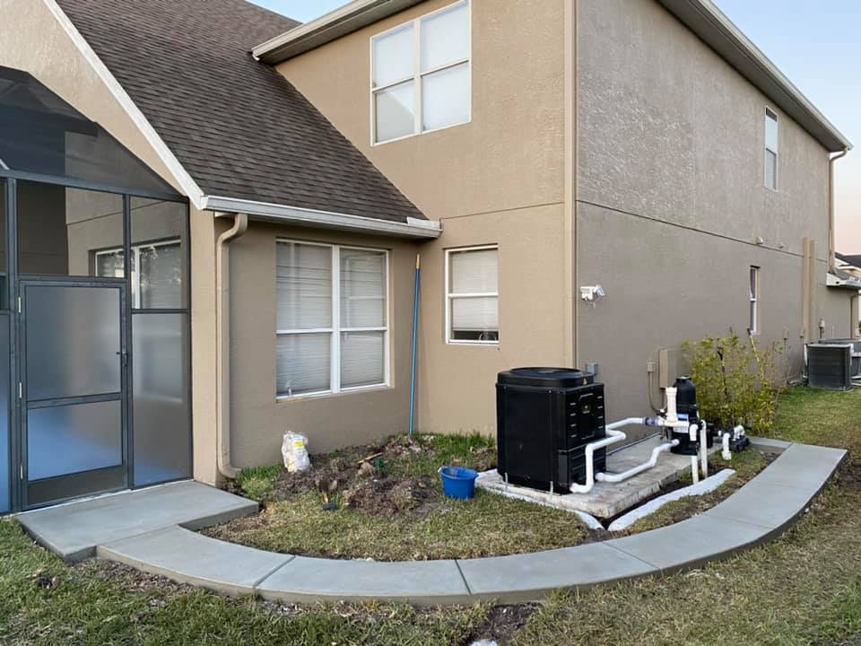 Exterior of a two-story house with a pool filter system and concrete walkway. Beige stucco walls and dark roof.