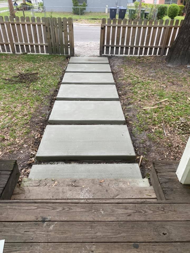 Concrete pathway leading from wooden porch steps to a picket fence.