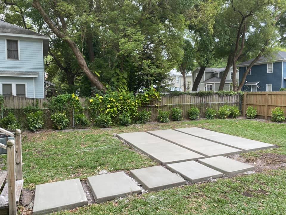 Concrete pavers form a patio and walkway in a grassy backyard, with houses and a wooden fence in the background.