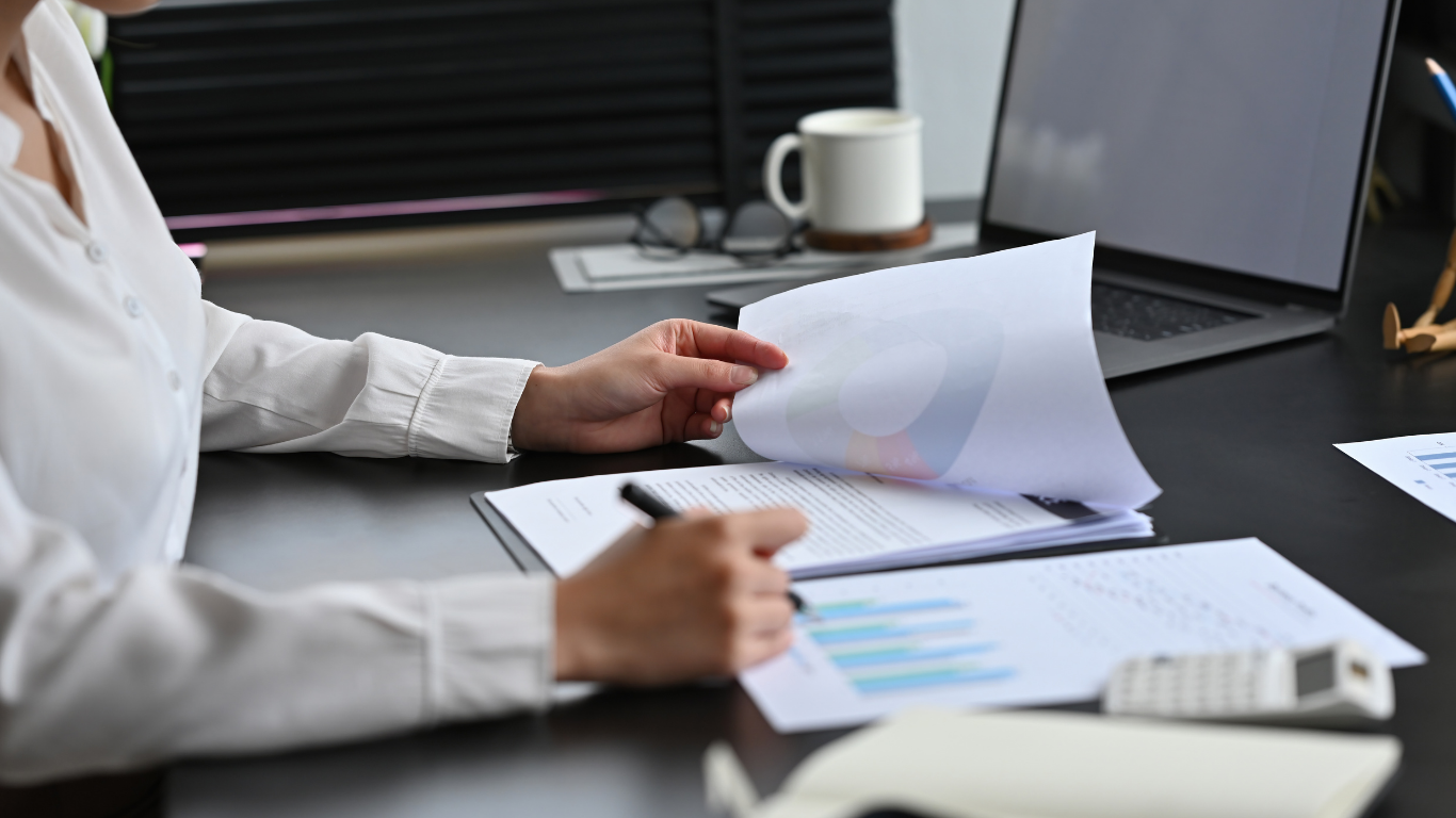 Man in suit examines documents at a desk, using a magnifying glass.