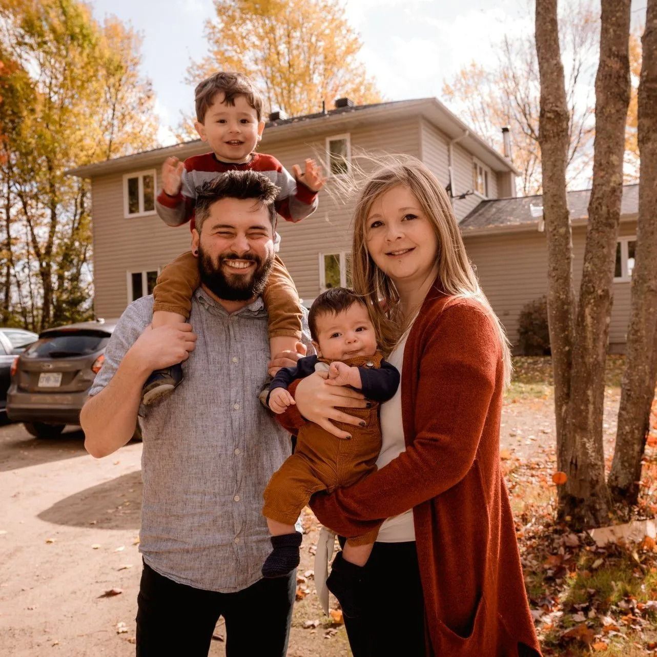 Family of four smiling outdoors in autumn, with two children on their parents' shoulders and in arms.