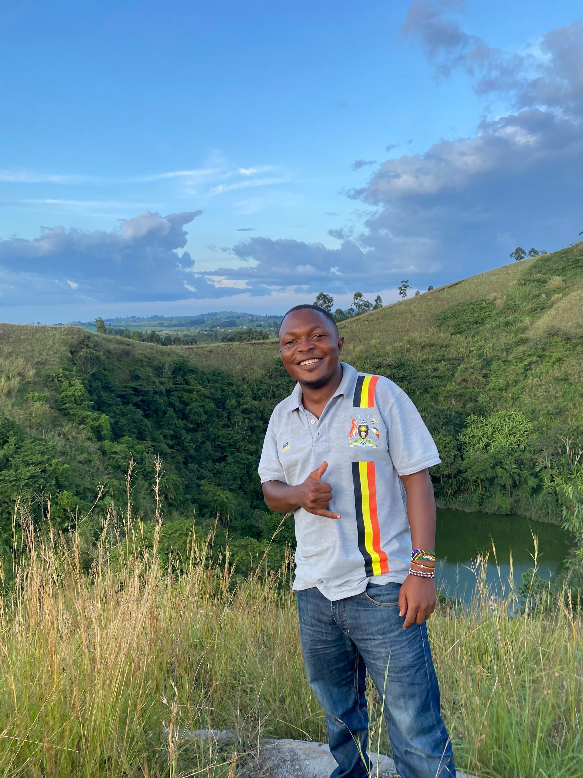 Man in grey shirt and jeans smiles, gives thumbs-up, standing near a lake in a grassy, hilly landscape under a blue sky.
