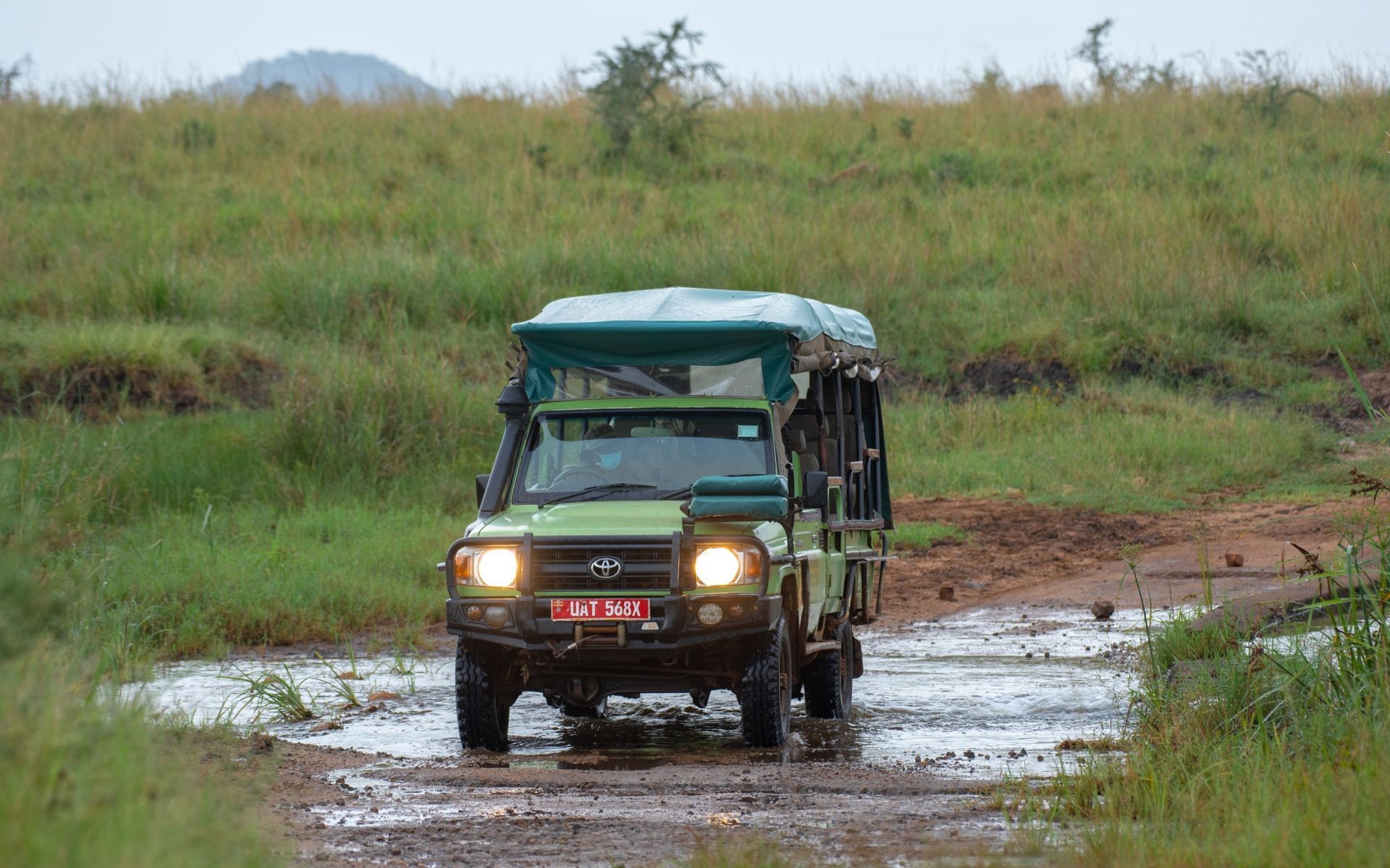 Green safari vehicle driving through a flooded dirt road in a grassy, hilly landscape.