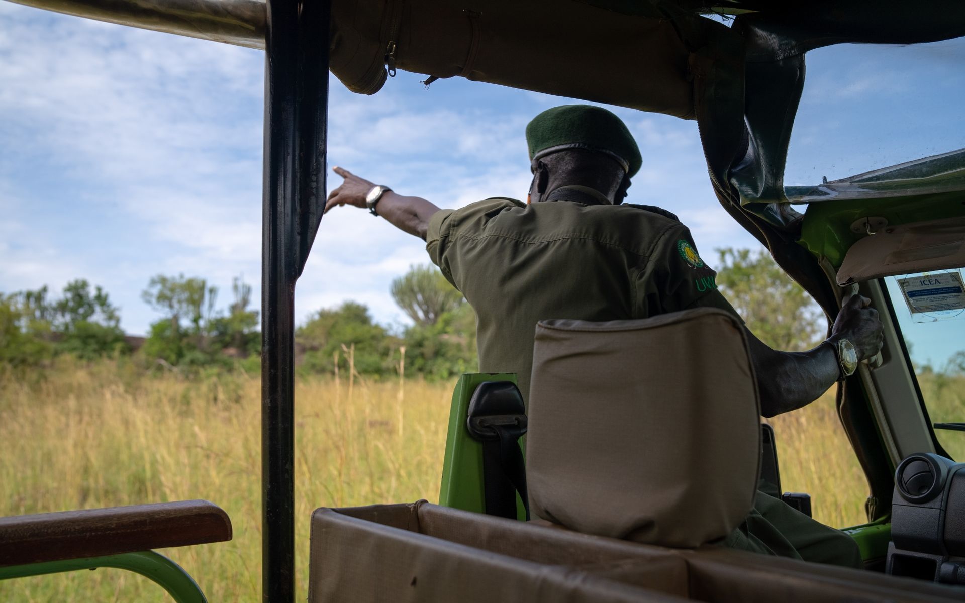 A park ranger in green uniform points toward the savanna from inside a safari vehicle.
