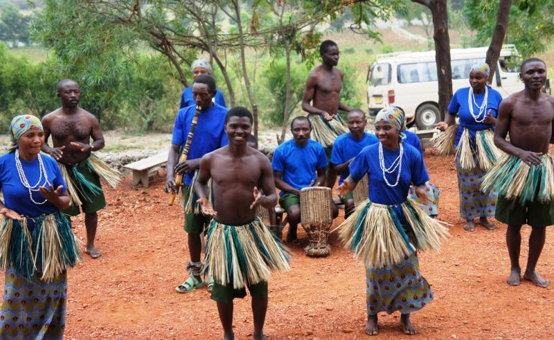 A group of African dancers, some topless, in blue tops and straw skirts, perform in a dirt clearing.