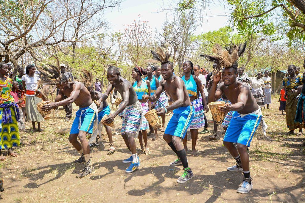 People dancing outdoors in blue shorts and tops, drums nearby, under a bright sky.
