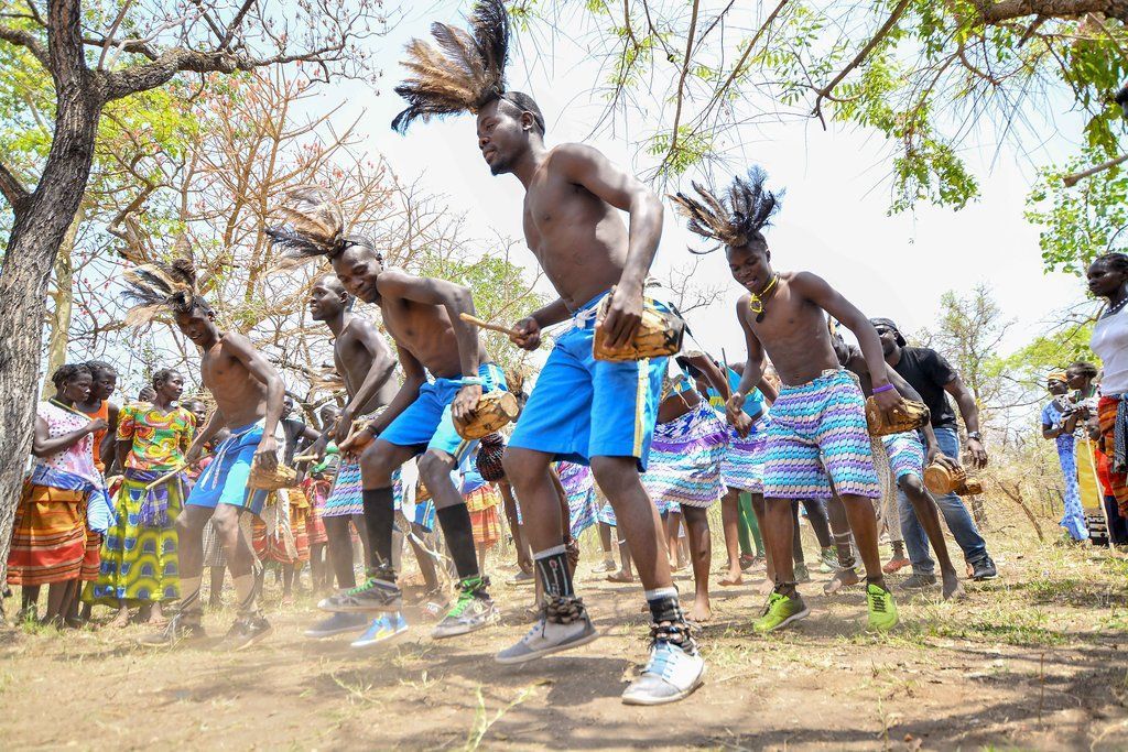 Men dancing outdoors, wearing feathered headdresses, blue shorts, and holding objects. Many people watching in background.
