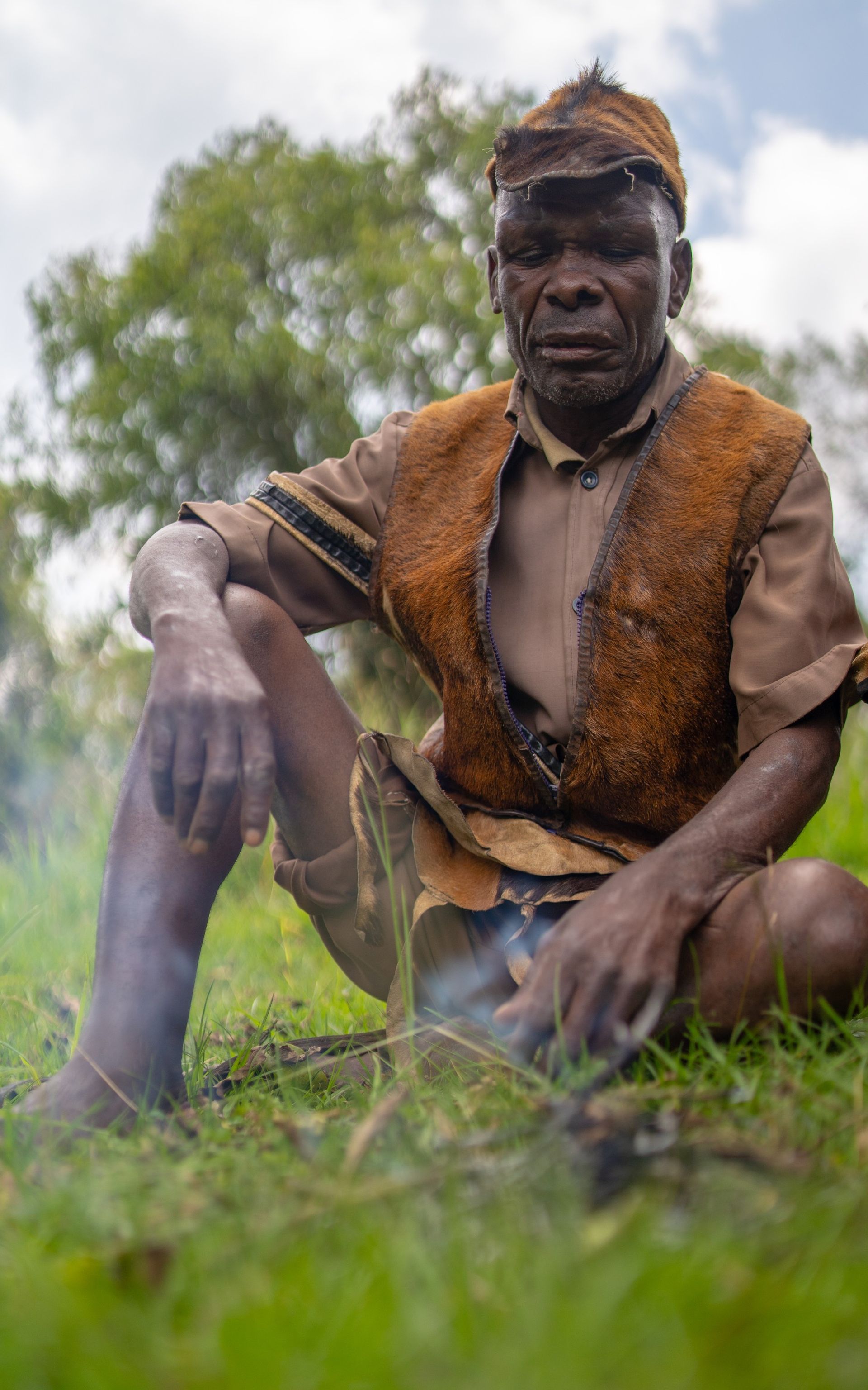 Indigenous man in leather vest sits by small fire in grassy field.