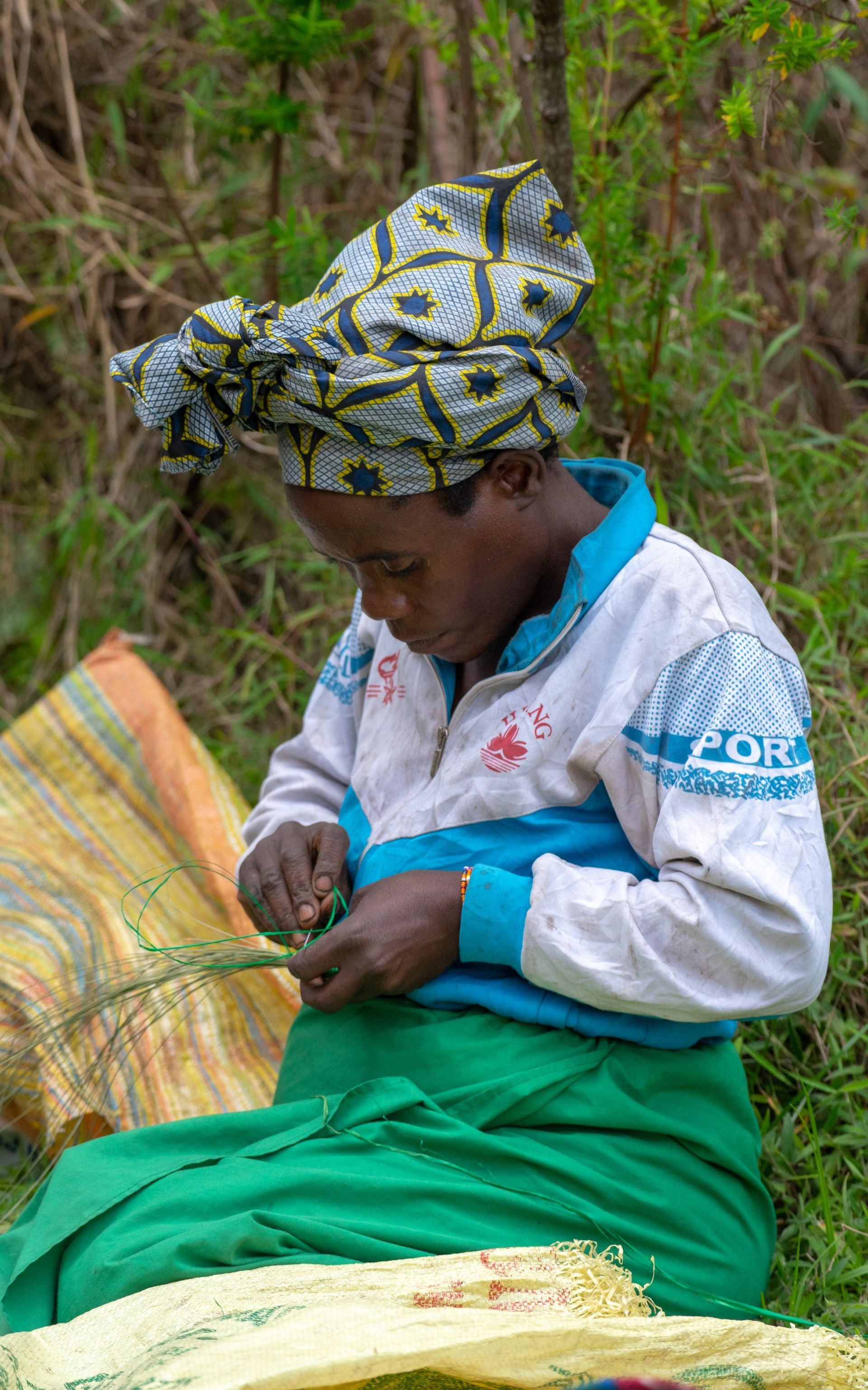Woman weaving grass, wearing a head wrap, blue and white shirt, and green skirt, seated outside.