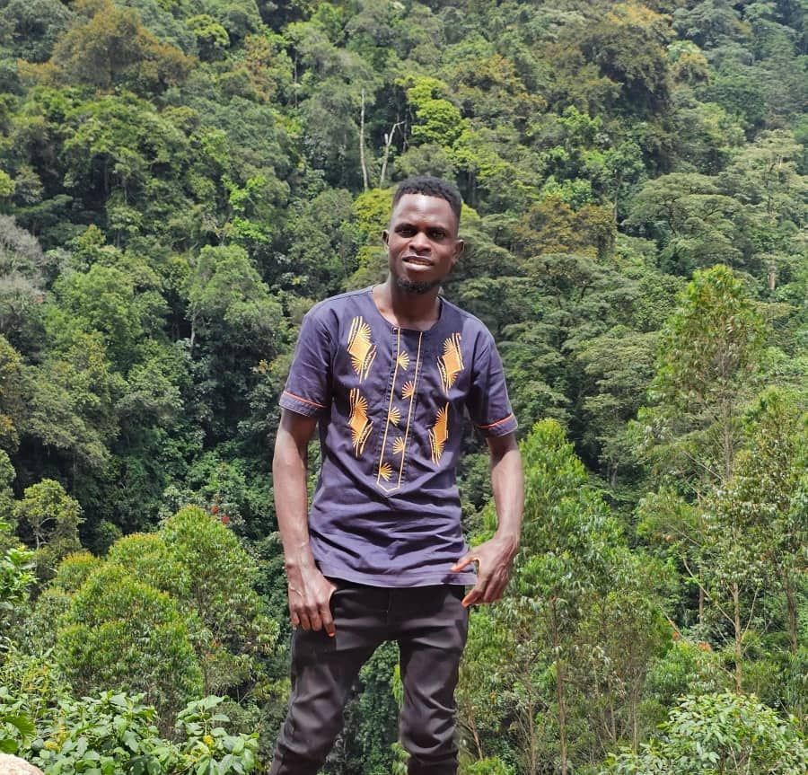 Man in blue shirt with gold designs stands before a lush green forest.