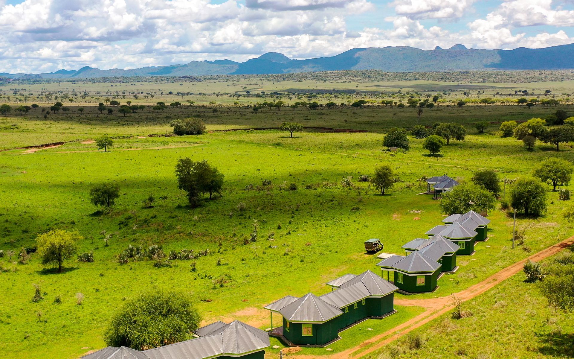 Green cabins in an African savanna, with distant mountains under a cloudy sky.