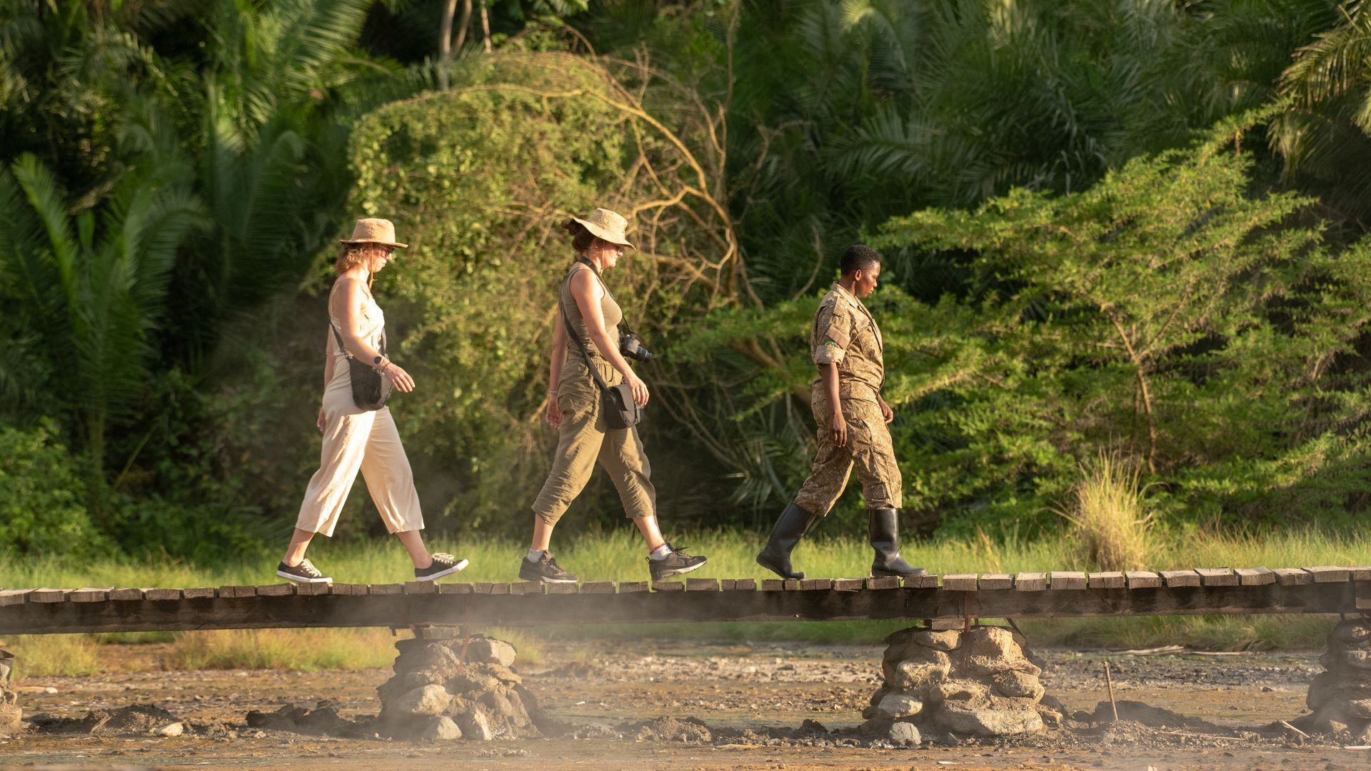 Three people wearing hats walk across a wooden bridge in a sunny outdoor setting with trees.