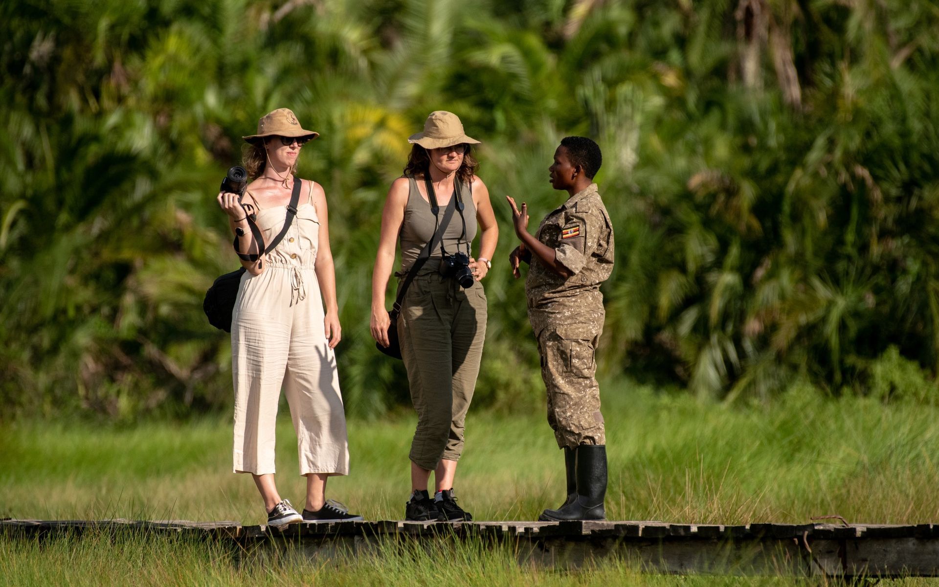 Three people on a boardwalk: two tourists in hats with a guide in uniform, gesturing. Lush green background.