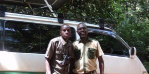 Two men in uniform stand next to a white van in front of a forest, smiling.