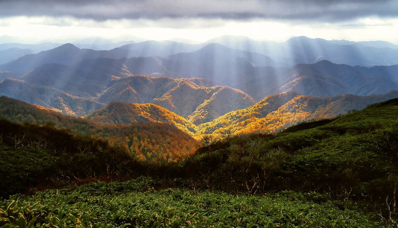 Sunlight streams through clouds, illuminating a mountain range covered in autumn foliage.