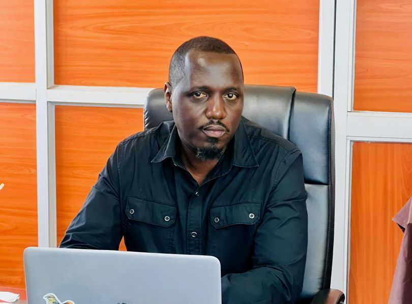 Man in black shirt sits at desk with laptop in front of an orange and white wall.