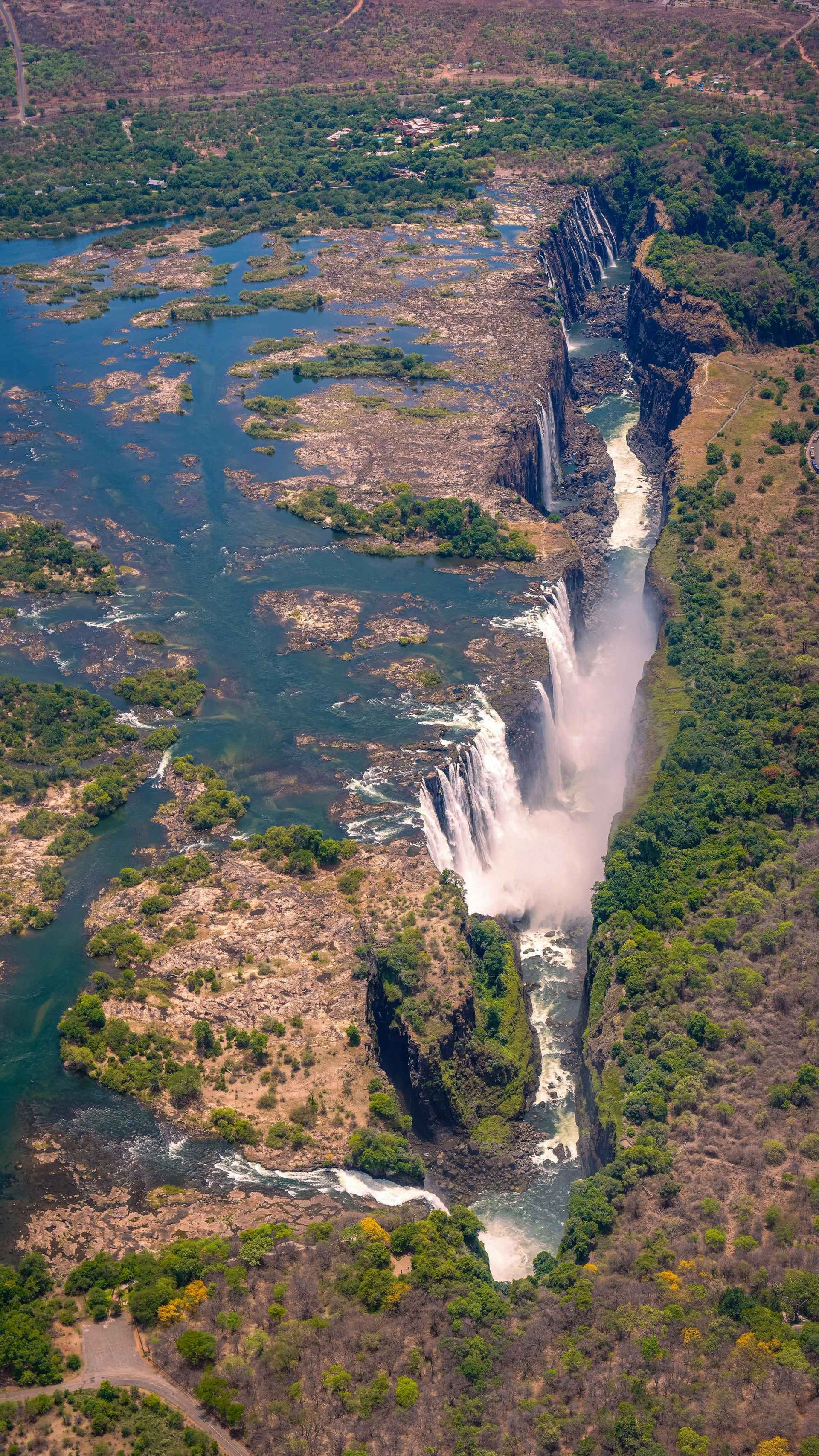 Aerial view of Victoria Falls cascading down into a deep gorge; river flows through green and brown landscape.